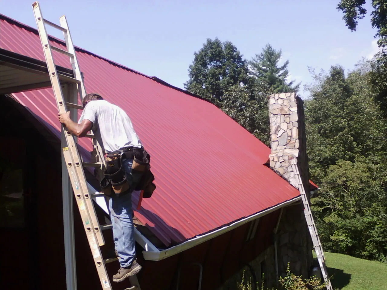Roofer on a ladder beside a red metal roof and stone chimney — Soffit & Fascia Repair by Roofing Brothers in Pinesdale